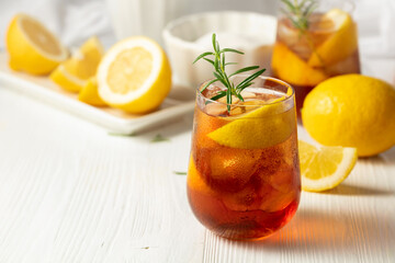 Iced tea or alcoholic cocktail with ice, rosemary and lemon slices on the white table.
