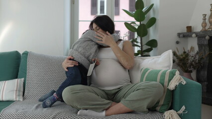 Child Embracing Pregnant Mother - Son Hugs Mom Expecting Unborn Brother on Home Couch in Late Stage...
