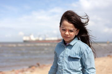A little girl in glasses stands on the sea beach. Large ship for transporting goods in the...
