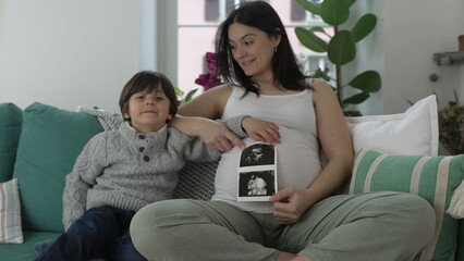 Happy Mother and Child Posing with Ultrasound Picture - Smiling at Camera with Unborn Baby Brother's Image, Seated on Home Couch in Third Trimester