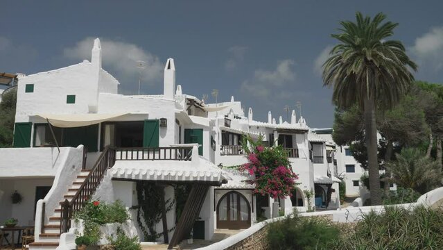 Mediterranean whitewashed houses with wooden windows, surrounded by Bougainvillea flowers and palm trees, picturesque travel destination in Binibeca Vell, Menorca, Spain