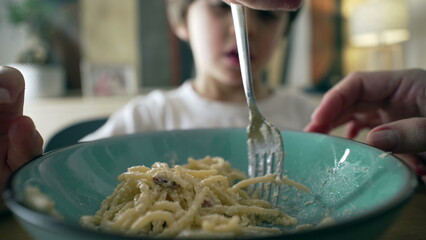 Close-up of fork spinning spaghetti on blue plate with little boy in blurred background, mother's...