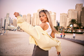 Stylish woman poses in a relaxed manner against a backdrop of skyscrapers on a city beach at dusk
