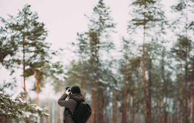 Young Man Backpacker With Photo Camera Taking Photo In Winter Snowy Forest. Active Hobby. Hiker Walking In Snowy Pinewood Forest.