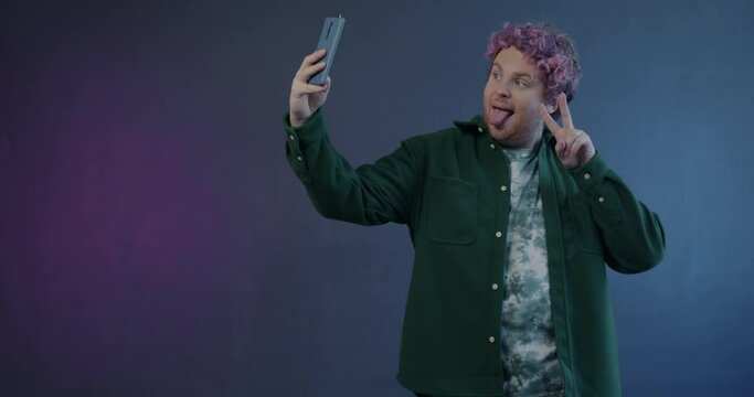 Slow motion portrait of playful young man taking selfie posing for smartphone camera having fun on purple background. Photo and modern gadget concept.