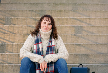 Relaxed woman sitting on urban steps, sporting a cozy sweater and scarf, a casual yet chic daytime look, concept: casual chic, comfort, city break - Powered by Adobe