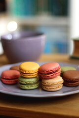 Purple plate filled with pastel macarons, cup of tea or coffee, vintage books and reading glasses on the table. Colorful bookcase in the background. Selective focus.