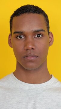 Vertical HD video. Closeup portrait of young african american man in white t-shirt looking serious at camera over yellow background. Youth and Gen Z People concept.