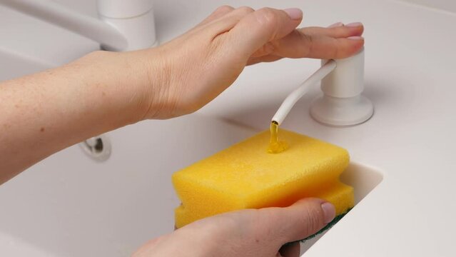Female hand press on built-in dispenser in the sink with detergent. Woman washing dishes by yellow sponge with soap in white kitchen. Housekeeping concept