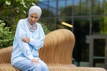 Muslim woman in hijab sitting outside, scratching her arm with a painful expression due to an...
