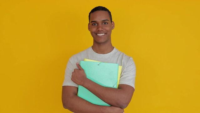 Confident portrait of smiling student boy holding academic file folder over yellow studio background. Education lifestyle concept.