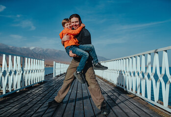 Happy father holds his son in his arms standing on a pier on mountains background