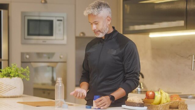 Maintaining Wellbeing in Modern Kitchen. Silver-Haired Mature Man Taking Vitamins or Supplements with Water Bottle. Focus on Personal Health and Daily Nutritional Routine in Homely Environment.