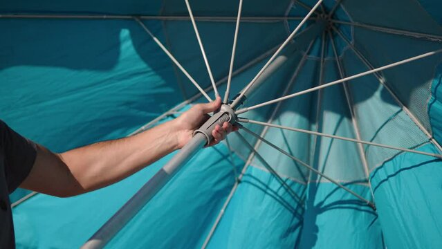 Man with his hands folds large beach umbrella with spokes on sea beach on sunny day, close up. Sun protection while relaxing on ocean coast. Tourist beach without amenities in season.