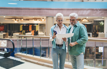 Elderly Caucasian couple discuss paperwork in a mall. Woman with phone and man with glasses show concern, implying financial or health matters. Light attire reflects a serene, spring setting.