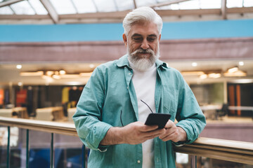 Smiling senior Caucasian man with beard, using smartphone, leaning on railing in mall. cheerful male embodies active retirement, comfortably engaging with modern technology in a public indoor space.