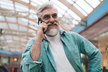 Joyful senior Caucasian man with beard, speaking on mobile phone in mall. man in his 60s, exudes happiness and comfort, hand on hip, engaging in pleasant conversation in bright, airy indoor setting.