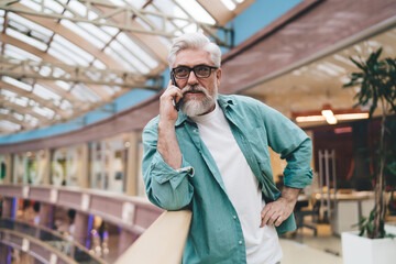 Confident elderly Caucasian man with white beard, speaking on phone, teal shirt, in a mall, hand on hip, pensive expression, standing indoors with daylight.