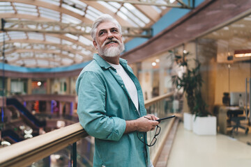 Joyful senior man with a white beard and teal shirt, holding eyeglasses, gazes upwards in a sunlit mall, embodying a serene retirement lifestyle.