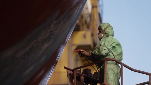 Worker sprays paint on ship hull in dry dock. Professional in protective gear applies red coating to vessel exterior. Maritime maintenance, shipbuilding industry scene. Dockyard labor.