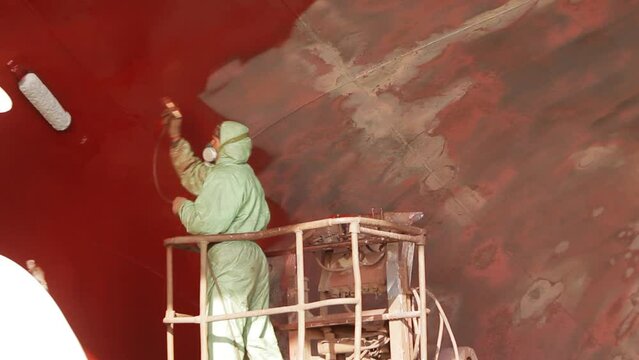 Worker in protective gear sprays red paint on ship hull in dry dock. Ship refurbishment, maintenance process in sequence, maritime industry. Professional with spray gun applies anti-corrosive coating