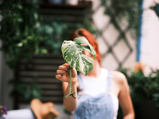 A red-haired woman standing in a garden, hides her eye behind a patterned leaf