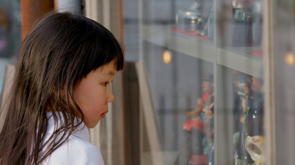 A girl is looking intently at toys though the shop window. - obrazy, fototapety, plakaty