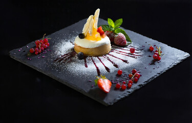 A cheesecake pastry with fruits and powdered sugar on a  black plate.