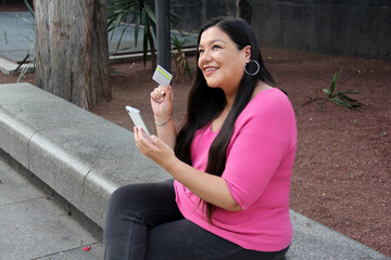 40-year-old Latina woman uses her cell phone and credit card to make payments, buy online, take...