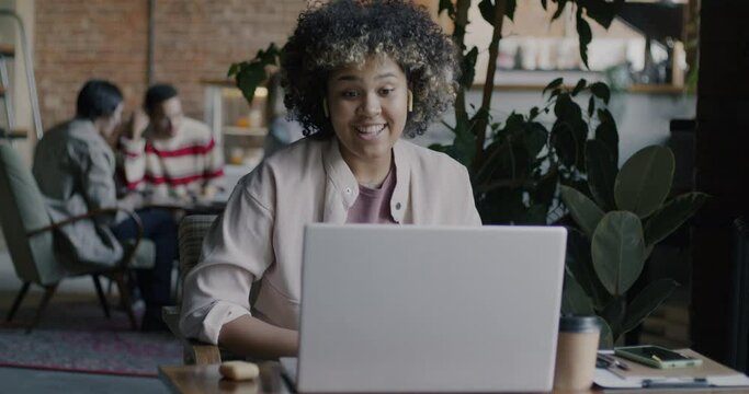 Female African American freelancer making online video call using laptop and wireless earphones in creative cafe. Freelance occupation and remote communication concept.