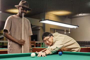 Portrait of two smiling friends playing pool together at billiards club with Asian man looking at camera copy space