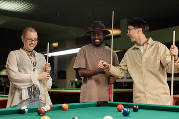 Waist up portrait of diverse group of friends enjoying game of pool together at billiards table and smiling cheerfully