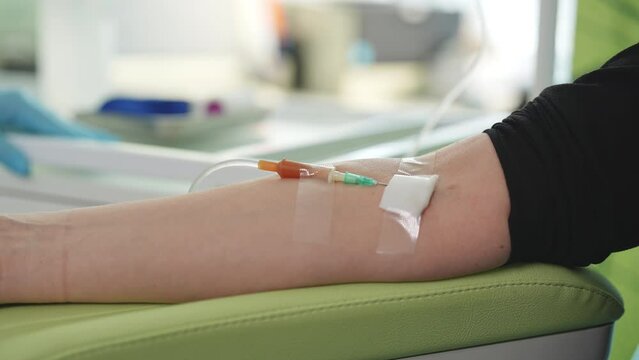 Close-up arms of unrecognizable female patient with IV catheter in vein receiving vitamin treatment in spa salon lying on comfy medical armchair. Healthcare, wellness and medicine concept, slowmo.
