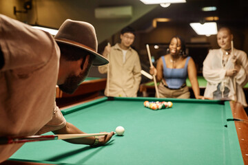 Over shoulder view of African American man playing pool and hitting ball with cue stick with diverse group of people in background copy space