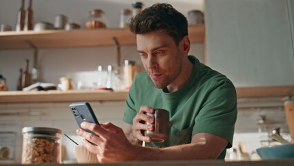 Positive guy browsing mobile phone having breakfast at home kitchen closeup