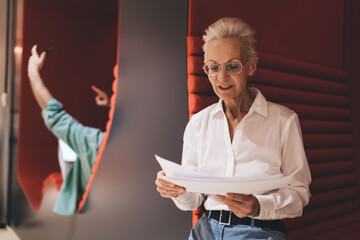 Focused senior woman reading documents, with a gesturing man in the foreground, in a dynamic office setting with red acoustic panels.
