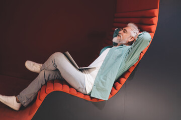 Relaxed senior Caucasian man with a white beard reclining peacefully on an orange lounger, holding a laptop, in a modern office lounge.