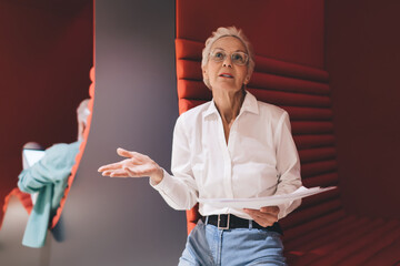 Communicative senior woman holding papers, gesturing in discussion, with a colleague's presence in a vibrant red office space