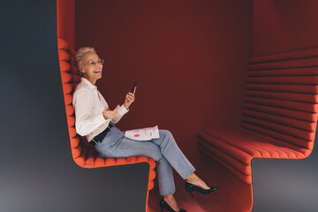 Cheerful senior businesswoman using a smartphone, sitting on a modern red bench with documents, enjoying a light moment in a creative office space.