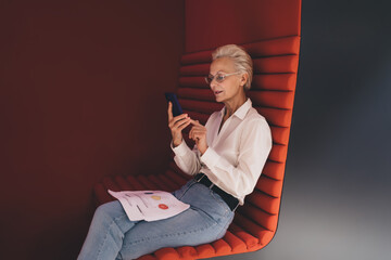 Senior businesswoman in a white shirt focused on her smartphone, with paperwork on her lap, seated on a red bench in a modern office