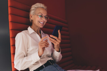 Joyful senior woman in white blouse texting on smartphone app seated on vibrant red acoustic office furniture, engaged and happy.