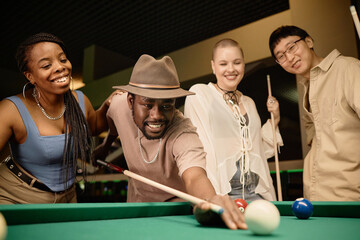 Multiethnic group of friends playing pool together with focus on smiling Black man hitting ball with cue stick copy space - 782450268