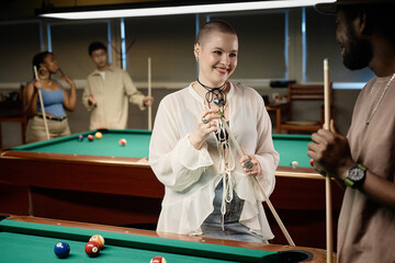 Waist up portrait of bald young woman chatting with friend and smiling while enjoying game of pool together in low light copy space