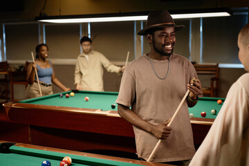 Waist up portrait of African American adult man chatting with friend and smiling while enjoying game of pool together in low light copy space - 782451866