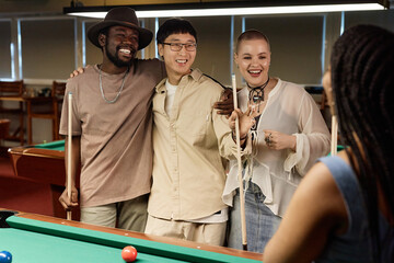 Waist up portrait of multiethnic group of friends chatting and smiling while enjoying game of pool together in low light