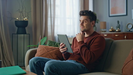 Relaxed customer looking tablet sitting couch living room. Handsome man swiping 