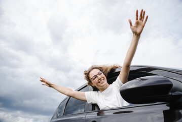 young beautiful woman in car looking forward, leaning out of car window to waist, waving hands happily, attractive caucasian woman in white t-shirt, traveling concept