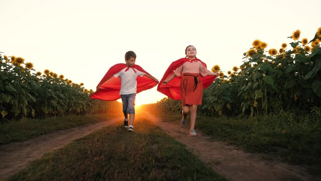 Happy children superhero in red cloak running image flying at sunset sunflower field. Boy and girl playing hero flight for protect planet fantasy imagination at sunrise sun light agriculture meadow
