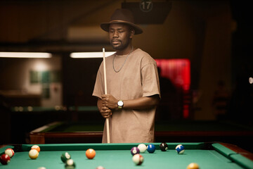 Waist up portrait of adult African American man wearing hat and holding cue stick while standing by table in pool club copy space