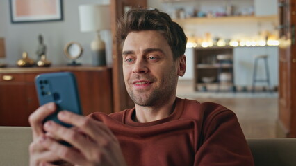 Smiling guy looking mobile phone screen on couch closeup. Man reading message 